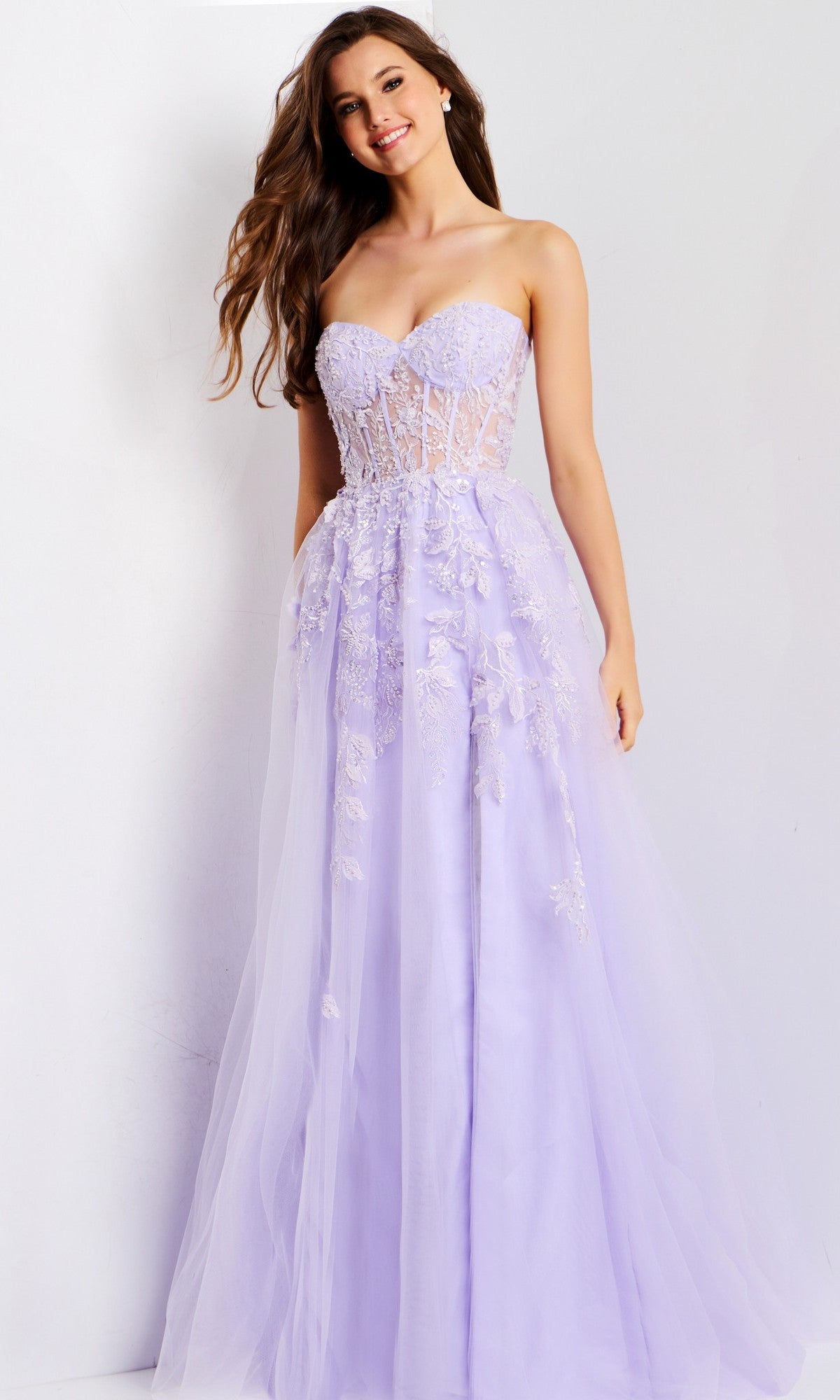 A woman wears the Jovani 49656 long prom dress in lilac, featuring a strapless sweetheart neckline, sheer lace-embroidered bodice, flowing tulle skirt, and delicate 3D floral appliques, standing against a plain light background.